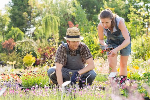 The image depicts a lush, well-maintained garden during daytime with vibrant flowering plants, including pink, purple, and yellow blossoms, surrounding a neatly trimmed lawn area. Two women are engaged in gardening activities; one is kneeling on the grass, dressed in casual gardening attire with a hat, tending to the flower beds with gardening gloves and tools, while the other is standing nearby, leaning forward slightly, holding a pair of pruning shears and wearing denim overalls. The garden features a background of mature trees and shrubs, providing a natural, shaded backdrop, with sunlight filtering through the foliage, creating a bright and inviting outdoor environment. The scene likely captures a moment of outdoor gardening, typical of local residential gardens in Dulwich, supporting professional gardening services offered by Landscapers Dulwich. The space appears organized with flower beds bordered by edging, and the overall setting emphasizes healthy plant growth and landscape care, consistent with lawn and garden maintenance in South London.