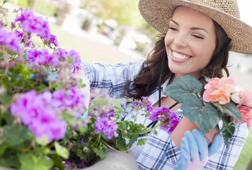 A woman wearing a wide-brimmed straw hat, a blue and white checked shirt, and gardening gloves is working in a garden filled with vibrant flowering plants. She is smiling while tending to potted plants that include purple, pink, and orange flowers, positioned on a garden table or planter. In the background, blurred garden elements such as trees, shrubs, and a well-maintained lawn are visible, suggesting a lush outdoor space typical of a residential garden in Dulwich, South London. The scene is illuminated by natural daylight, indicating pleasant weather conditions, and the garden appears to be well-organized with a mix of flower beds and greenery, supporting local gardening and landscaping services like those offered by Landers Dulwich to enhance outdoor aesthetics.