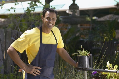 A smiling male gardener wearing a yellow T-shirt and navy apron stands in a lush garden, holding a small black pot filled with green plants. The garden features a mix of well-maintained grass, various shrubs, and flowering plants, with wooden fencing and a garden shed visible in the background. The scene is illuminated by natural daylight under a partly cloudy sky, with some patio furniture and garden ornaments in the distance, suggesting a landscaped outdoor space typical of residential areas in Dulwich. The gardener appears to be involved in planting or garden maintenance, demonstrating professional outdoor gardening services offered by Landscapers Dulwich in the local South London area, with a focus on lawn care and landscaping elements.