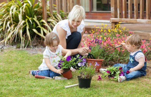 A woman with blonde hair, wearing a white top, is kneeling on a neatly maintained grassy lawn in a garden in Dulwich, South London, engaging with two young children, both girls, who are sitting on the lawn. The children are dressed in casual clothing and are involved in planting or arranging vibrant purple, pink, and yellow flowers in small garden pots and flower beds. The garden features a lush green lawn, bordered by a flower bed filled with colorful flowers, and is situated adjacent to a wooden deck area with steps leading to a home in the background. Surrounding plants include ornamental grasses and shrubs, adding to the lush, welcoming outdoor space. The scene is set in daytime under natural light, suggesting a clear, pleasant weather suitable for outdoor gardening activities. This image evokes professional gardening and outdoor maintenance services such as planting, lawn care, and garden tidying provided by Dulwich-based landscapers like Landscapers Dulwich, emphasizing a nurturing environment suited for family and outdoor leisure.