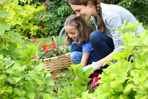 A woman and a young girl are gardening together in a lush, well-maintained backyard garden, surrounded by dense greenery, flowering plants, and shrubs. The girl is crouched down, holding a woven basket filled with small red flowers, while the woman kneels beside her, guiding and supporting her gardening activity. The garden features a mix of vibrant green foliage, with some plants showing broad leaves and others with delicate, smaller leaves. The ground is covered with grass, and the scene is illuminated by natural daylight, suggesting a clear, pleasant day. In the background, there are various garden elements such as flower beds, possibly bordered with soil or mulch, and a hint of garden path or paving stones. This outdoor space exemplifies a tidy, healthy garden environment suitable for family gardening activities, reflecting professional landscaping and gardening care that Landscapers Dulwich offers to enhance outdoor spaces in the Dulwich area.