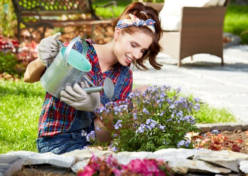 A woman with a cheerful expression, wearing a plaid shirt and protective gardening gloves, is kneeling on a garden lawn while watering a small flowering shrub with light purple blooms using a metal watering can. She has a colorful headband holding back her hair and appears to be engaged in garden maintenance. In the background, there is a paved patio area with outdoor furniture, including a white table and brown chairs, set in a lush, green outdoor space. The garden features a well-maintained grass lawn, edged with a stone border, along with typical garden plants and mature trees, suggesting a suburban area possibly in Dulwich. The scene is illuminated by natural daylight with a slightly overcast sky, emphasizing natural tones of the garden elements. This image subtly reflects gardening and outdoor maintenance themes relevant to local landscaping services provided by Landscapers Dulwich, supporting content related to garden upkeep and flower bed care in the Dulwich area.