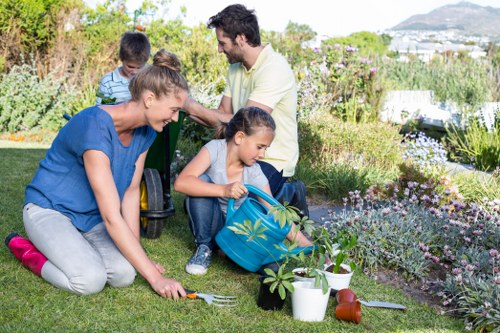 A family gardening together in a well-maintained backyard garden, featuring a lush green lawn with a mix of flowering plants and shrubs along the borders. In the foreground, a woman is kneeling on the grass, planting or tending to a small plant. Nearby, a young girl is sitting on the ground, handling garden tools and a watering can, with a garden fork placed in the grass. In the background, a man and two children are seated on a garden bench, actively engaged in planting or discussing their gardening tasks, surrounded by flower beds with vibrant blossoms and foliage, as well as neatly trimmed hedges and trees. The outdoor space is bathed in natural daylight, with clear weather and a scenic view of distant hills or countryside visible beyond the garden. This scene showcases an outdoor space suitable for gardening activities and outdoor family leisure, supported by the garden landscape design and maintenance typical of outdoor environments managed by professional gardening services in Dulwich or South London.