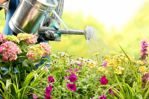 The image depicts a close-up view of a gardener's hands wearing gardening gloves, using a metal watering can to water a vibrant flower bed in a garden. The flower bed is densely planted with a variety of flowering plants including pink, yellow, purple, and white blooms, with some taller green foliage and grass blades visible in the foreground. In the background, there are lush green trees and shrubs, indicating a well-maintained outdoor space on a bright, sunny day with clear weather. The garden features a mix of flowering plants and dense foliage, with natural grass areas and a defined flower bed border. The scene highlights outdoor gardening activities typical of professional landscaping services offered by companies like Landscapers Dulwich, serving local areas such as Dulwich, with a focus on healthy plant growth and garden maintenance.