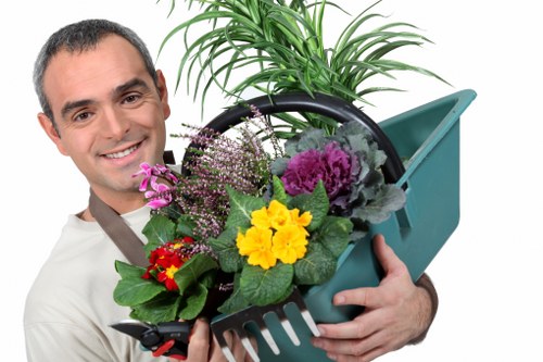 A man with short dark hair and a friendly smile holds a teal plastic gardening trowel and a matching hand rake, filled with colorful flowering plants, while standing outdoors in a garden setting. The garden features a well-maintained lawn with lush, dense, bright green grass in the foreground, bordered by a neat flower bed with various flowering plants displaying yellow, purple, and red blooms. In the background, there are several small trees and shrubs, some with freshly pruned branches, under bright natural daylight conditions. The scene suggests a tidy, landscaped backyard or front garden typical of residential properties in Dulwich, with a clear emphasis on healthy lawn care and plant maintenance, aligning with services provided by Landscapers Dulwich. The overall environment conveys a sense of professional garden upkeep suitable for outdoor landscaping and gardening work in south-east London postcode areas.