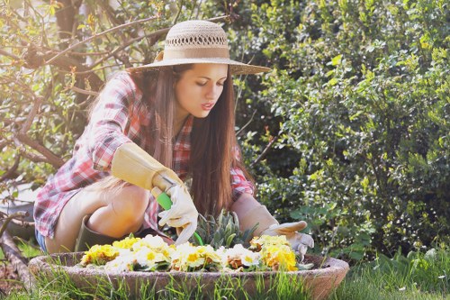 A young woman in a plaid shirt, sun hat, and gardening gloves is kneeling on a well-maintained lawn in a back garden. She is tending to a flower bed filled with yellow, purple, and white blooms, surrounded by lush green foliage and shrubbery. The garden features a neatly cut grass area, bordered by a low, curved edge, with mature trees and dense bushes creating a natural backdrop. Sunlight filters through the leaves, casting warm highlights on her focused expression as she carefully arranges or inspects the flowers. The scene reflects a peaceful outdoor environment suitable for gardening and landscaping services in Dulwich, highlighting the care needed for flower beds and lawn areas that Landscapers Dulwich may assist with in maintaining or improving outdoor spaces.