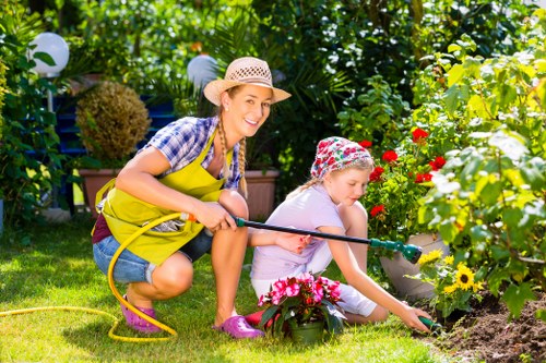 A woman and a young girl are gardening together in a lush outdoor space, with the woman holding a garden hose and smiling towards the camera while kneeling on a well-maintained grassy lawn. The girl is focused on planting or tending to a flower bed filled with colorful blooms, including red, yellow, and pink flowers, and is wearing a floral hat and casual clothing. The garden surrounding them is dense with green foliage, including a neatly trimmed hedge, shrubbery, and trees that provide natural shade. In the background, decorative garden lights and potted plants enhance the vibrant atmosphere. The scene takes place on a sunny day with bright natural lighting, reflecting a peaceful and productive outdoor environment typical of gardening and landscaping services in Dulwich and the surrounding South London area, emphasizing a well-organized, welcoming garden space.