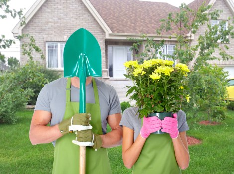 In a suburban garden with a well-maintained lawn, two individuals are engaged in outdoor gardening activities during daylight. The person on the left wears a green gardening apron, a grey t-shirt, and a teal protective face shield, holding a shovel with a teal blade, possibly for planting or soil work. The person on the right is dressed in a grey t-shirt and pink gardening gloves, holding a pink pot filled with vibrant yellow flowers, likely chrysanthemums or similar, close to their face. The garden features a neatly trimmed lawn with lush green grass, bordered by flower beds and small shrubs, and a backdrop of a residential house constructed with light grey bricks and a pitched roof, partially obscured by a leafy tree with light green foliage. The natural lighting suggests a clear day with some sunlight, providing a bright and inviting outdoor environment. This scene highlights professional gardening and landscaping practices suitable for residential gardens in Dulwich or nearby areas, reflecting the services offered by Landscapers Dulwich in maintaining and enhancing outdoor spaces.