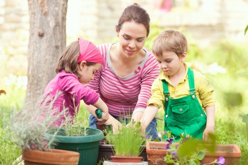 A woman with dark hair tied back, dressed in a pink and white striped shirt, is tending to potted plants in a garden alongside two children; a girl wearing a pink headband and purple top, and a boy in a yellow shirt with green overalls. They are gathered around a table with various flower pots containing different plants, with some pots made of terracotta and others of plastic, all placed on a wooden surface. The garden setting features lush green grass, a large tree trunk in the background, and a flower border with white blossoms, indicating a well-maintained outdoor space suitable for gardening activities. The scene is lit by natural daylight, suggesting a pleasant weather condition. This image illustrates outdoor gardening activities typical of residential gardens in Dulwich or similar local areas, with a focus on planting and nurturing garden plants, supporting professional gardening and landscaping services offered by Landscapers Dulwich.