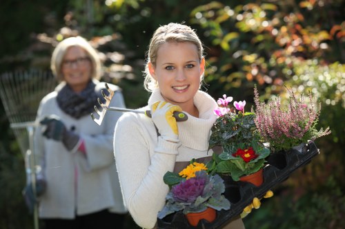 A young woman with blonde hair, wearing a cream sweater and gardening gloves, holds a black tray filled with colorful flowering plants, including red, yellow, purple, and pink blooms, in a well-maintained outdoor garden. Behind her, an older woman with glasses, a scarf, and a light jacket is slightly out of focus, holding gardening tools, with lush green shrubs, a variety of trees, and a flower bed visible in the background. The garden is bathed in natural sunlight, highlighting the vibrant colours and healthy soil, suggesting active gardening or planting work typical of a residential outdoor space in Dulwich, with neatly trimmed lawn edges and paved pathways visible. The environment appears calm, sunny, and suitable for gardening and outdoor maintenance activities, aligning with landscaping services offered by Landscapers Dulwich, based near the postcode area in South London.