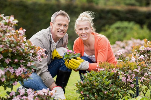 A smiling man and woman are kneeling in a well-maintained garden with a lush green lawn, flower beds filled with pink and white blossoms, and neatly trimmed shrubs. The man has short grey hair and is wearing a light grey shirt and gardening gloves, holding a small plant. The woman has blonde hair tied back, wearing an orange top and yellow gloves, also holding the plant. The background shows a hedge and trees, indicating a peaceful outdoor space likely in Dulwich, with natural daylight highlighting the vibrant colors of the flowers and greenery. This scene reflects outdoor gardening activity and maintenance, typical of professional landscaping and planting services provided by companies like Landscapers Dulwich, which specializes in creating and caring for beautiful garden environments in the local area. The overall environment suggests a pleasant, well-organized residential garden ready for further planting or improvement. The natural weather conditions appear bright and clear, ideal for gardening work in a suburban setting.